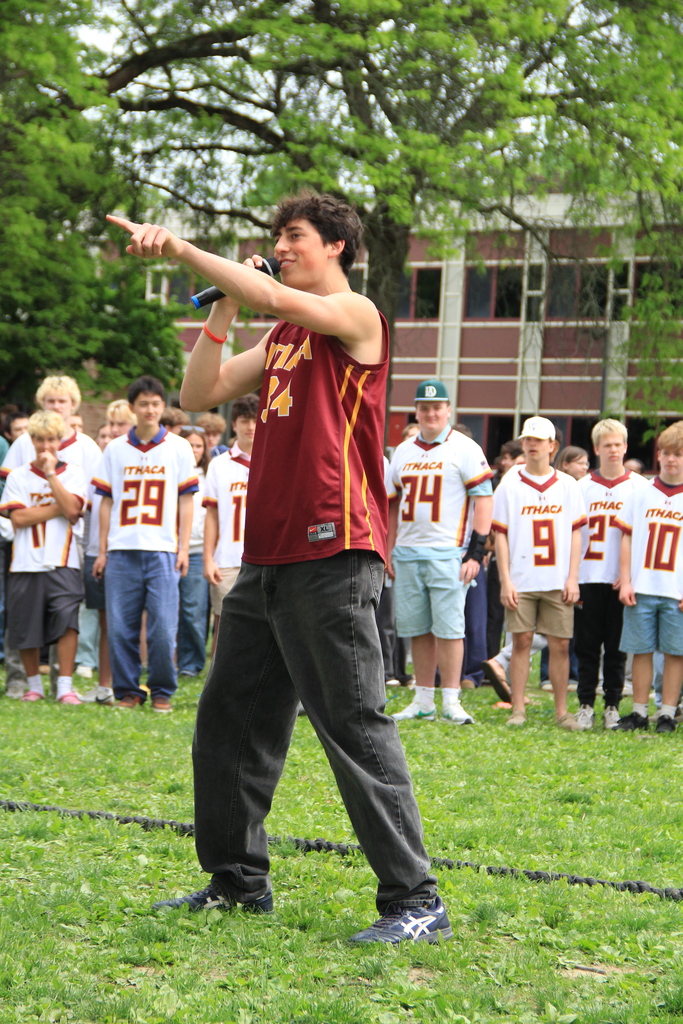 A person with a microphone in a maroon jersey performs in front of a group of children wearing numbered shirts.