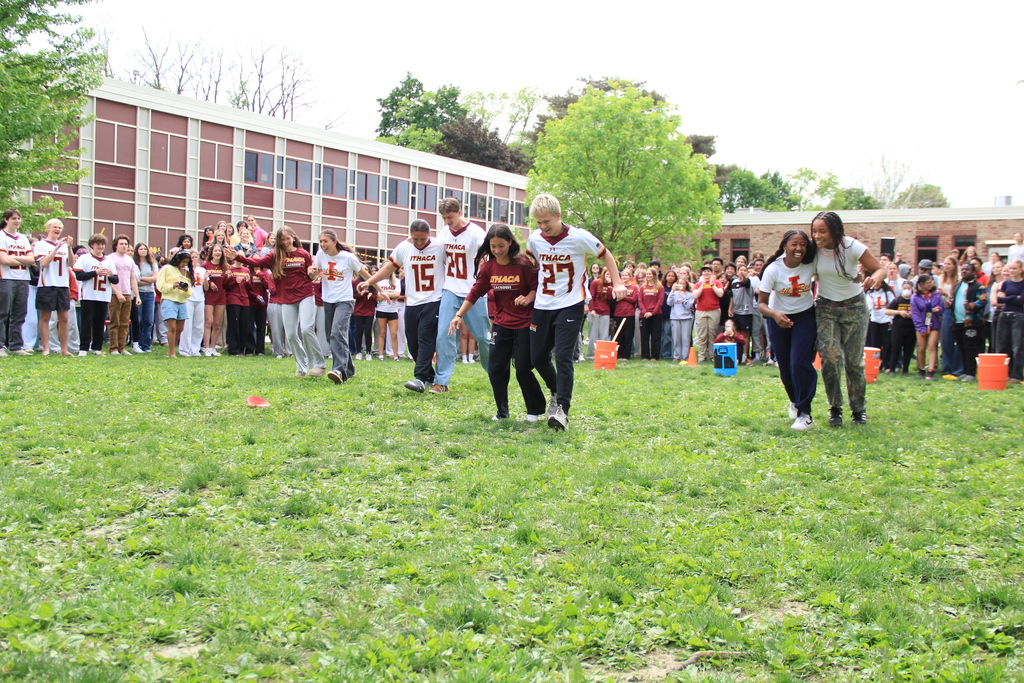 Group of people running across a grassy field with numbered jerseys, spectators standing behind them in front of a building.