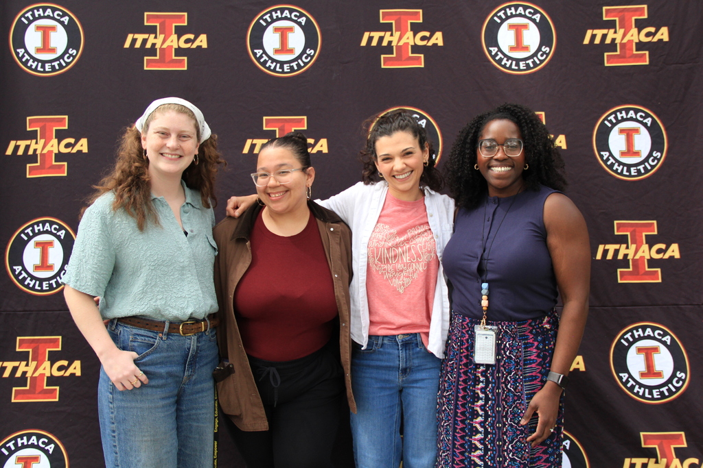 Four women pose together in front of a wall with logos and lettering reading "ITHACA" and "ATHLETICS".