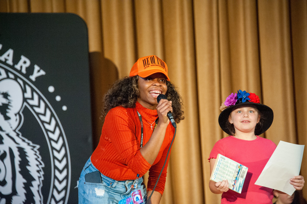 Woman with orange cap and orange top speaks into microphone, standing beside a girl holding papers.