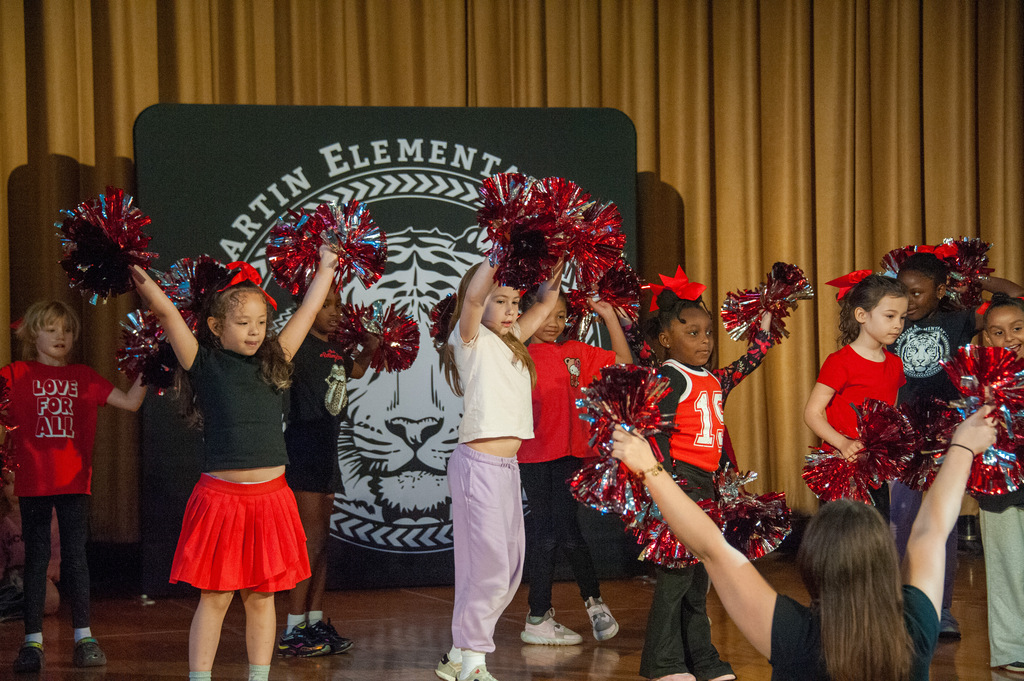 Group of children on a stage performing with pom-poms in front of a sign.