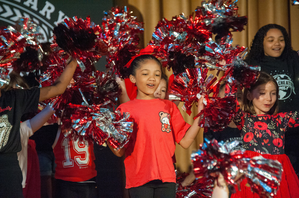 Children in red and black uniforms hold silver and red pompoms, performing on a stage.