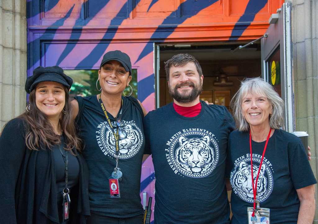 Four people wearing matching shirts with tiger logos, standing together in front of a colorful building.