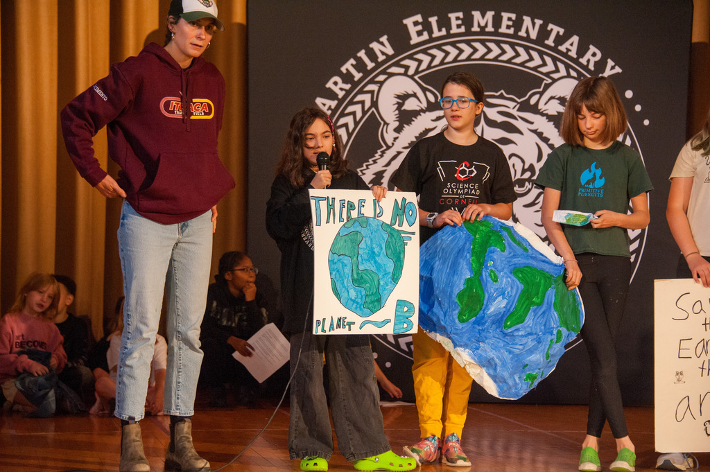 Four kids stand on stage, holding a large Earth poster and signs. A person stands behind them.