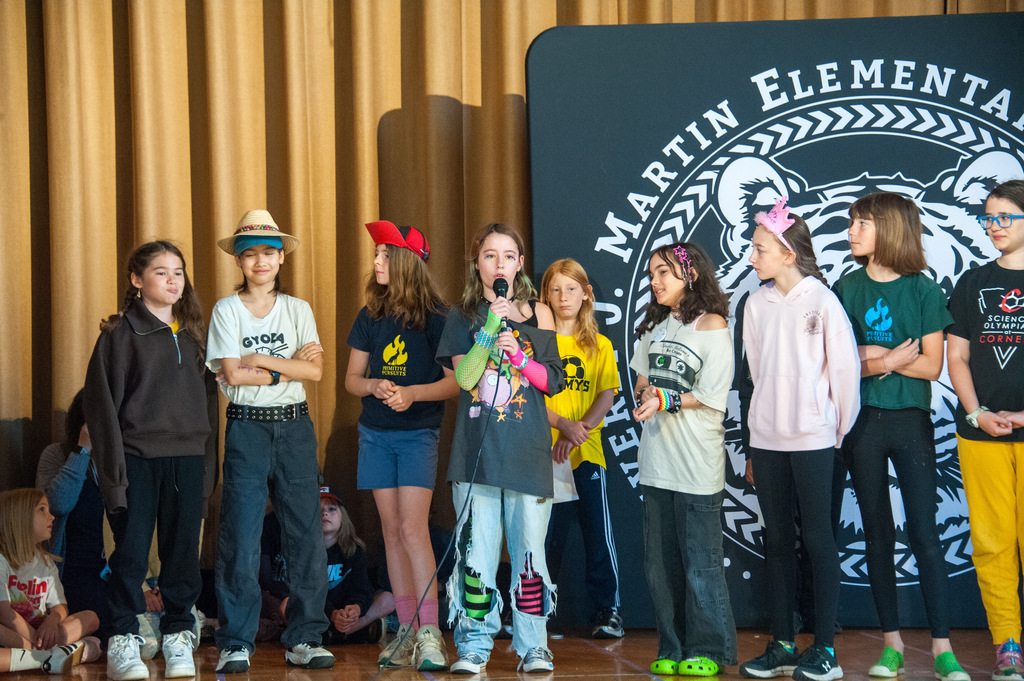 A group of students stands on a stage with one holding a microphone. They are wearing casual clothes and standing before a backdrop with the Martin Elementary logo.