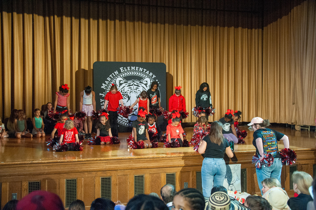Children wearing red shirts and red hats perform on a stage. A banner with a tiger logo is behind them.