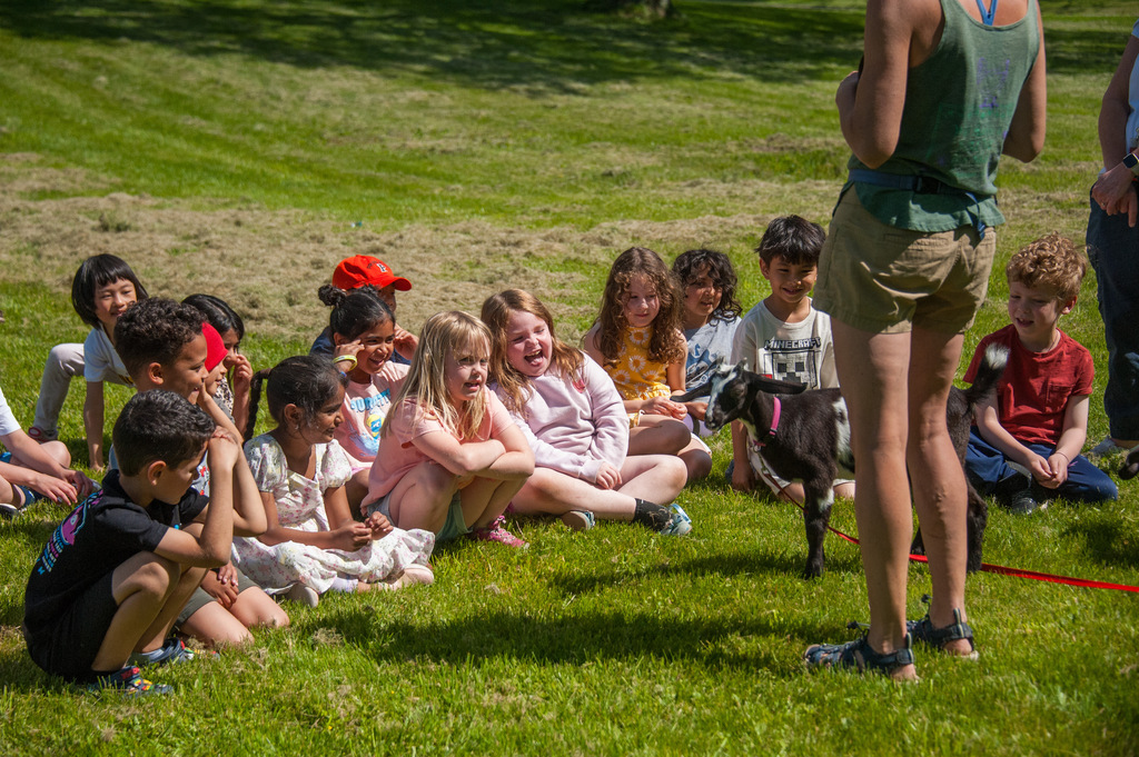 A group of children seated in a circle with a goat, a woman stands nearby. Green grass.