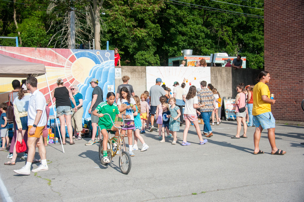 A group of people stands in an outdoor area. Some are wearing hats, sneakers, and sandals. One person rides a bicycle.