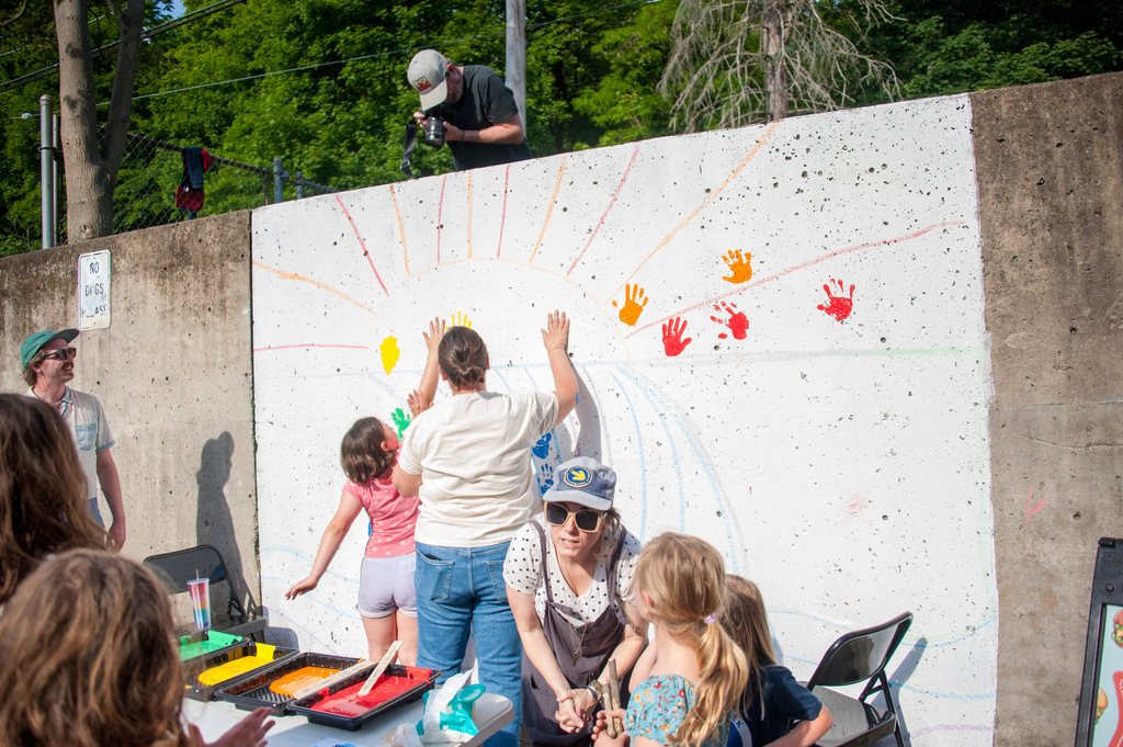 A group of adults and children standing around a mural on a wall, with one person painting.