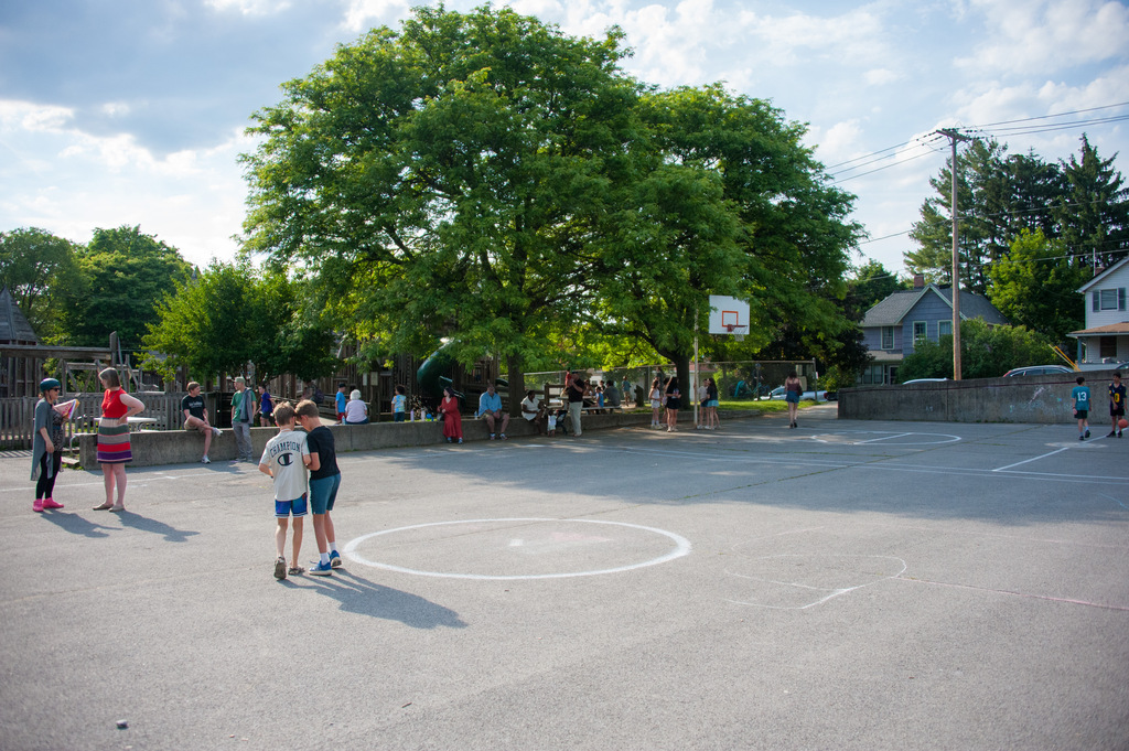 Several people stand in an open, paved area with a basketball court. A tree stands in the center.