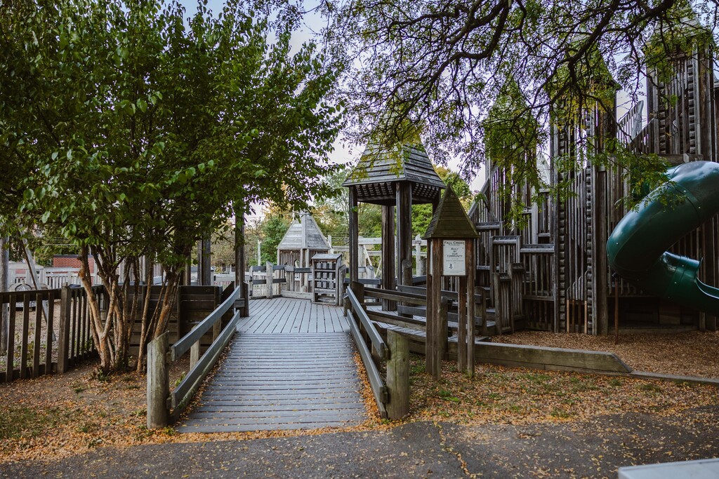 A wooden bridge leads to a wooden play area with a green slide, surrounded by trees.