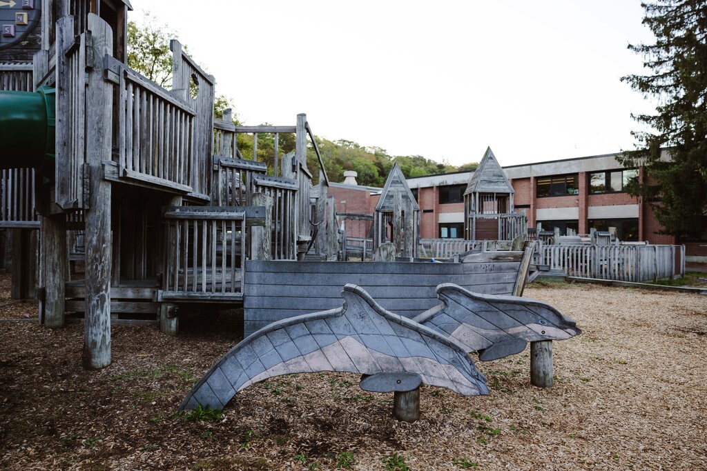 A children's outdoor play area featuring a wooden structure, dolphin figures, and a slide, with a building in the background.