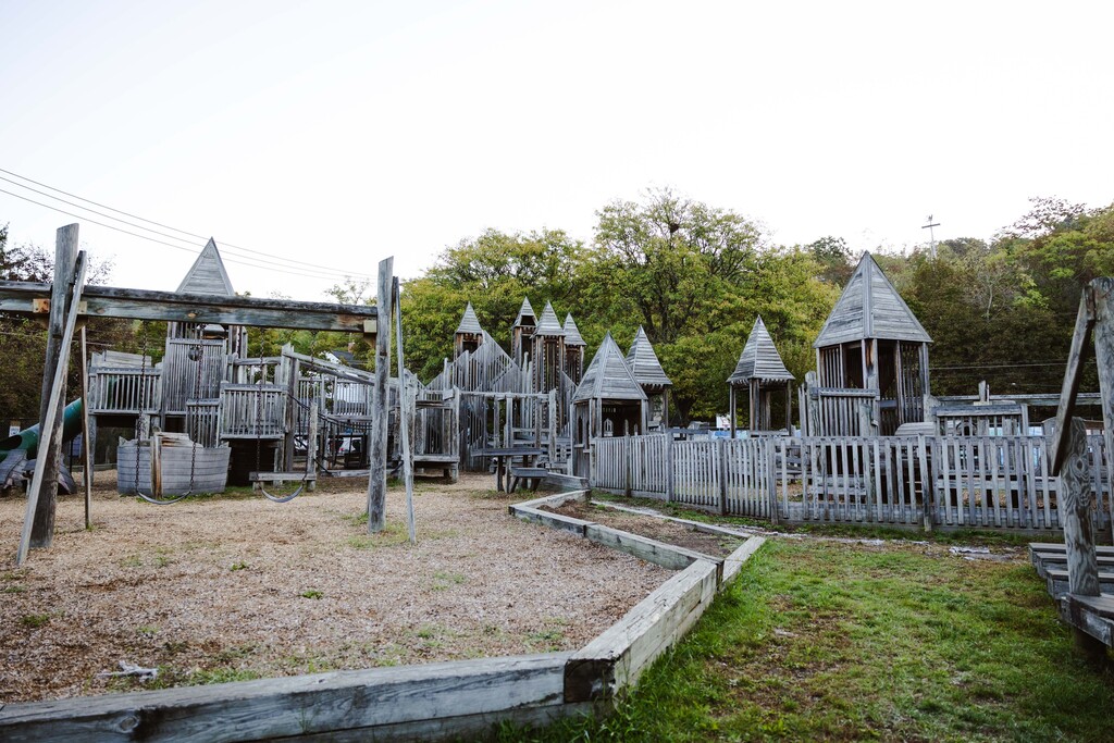A children's play area with wooden structures, a swing, and a small fence, set in a grassy area with trees in the background.