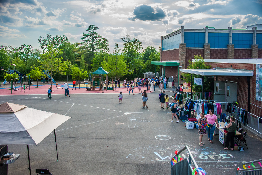 An outdoor area with people walking, a building with a tent, a playground, trees, and chalk drawings on the ground.