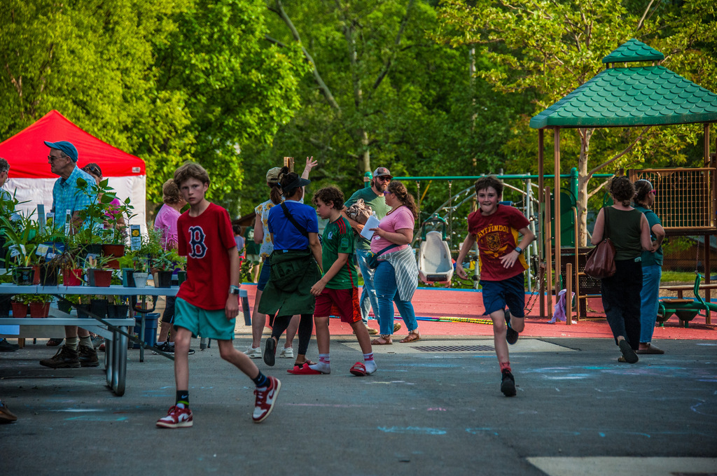 Children run and play in a park. Some wear red shirts, and others wear green. A gazebo and market area are in the background.