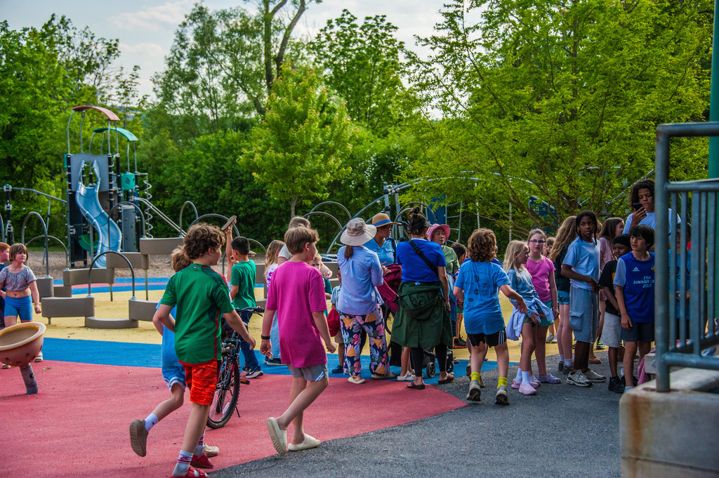 Kids and adults gather in a playground with trees in the background and a slide visible.
