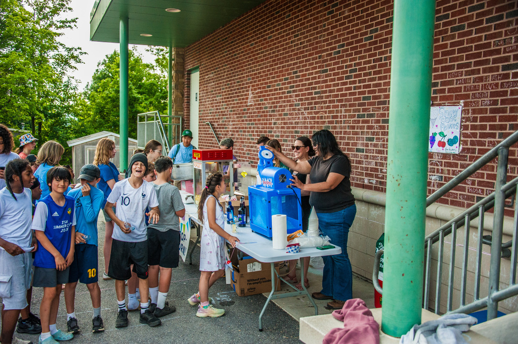 People, including children, gather near a building. A woman stands behind a table with a blue machine, and several children are waiting.