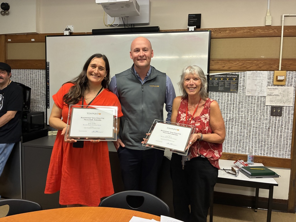 Three adults, two women and a man, hold framed certificates and stand together in a classroom.