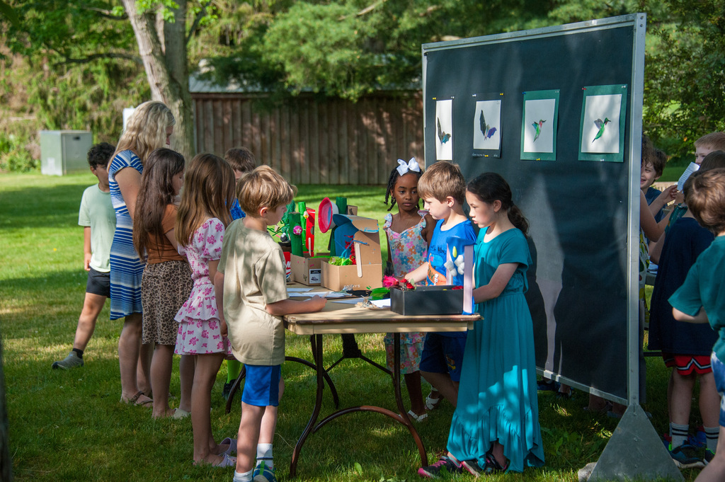 Kids and adults stand around a table with various crafts. A black board with artwork is behind them.