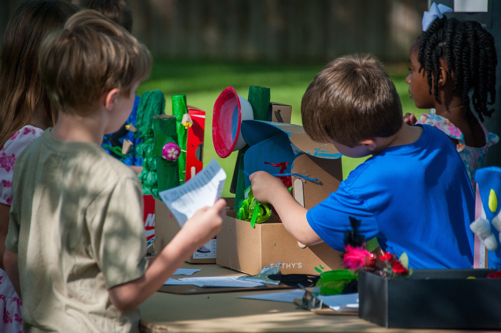 Children gather around a table, focusing on a cardboard craft. One child holds paper while another manipulates a colorful object.