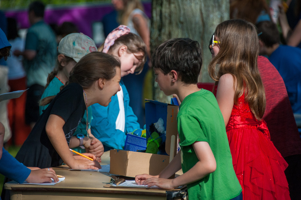 Children stand around a table with papers and pens, some signing. A tree trunk is visible in the background.
