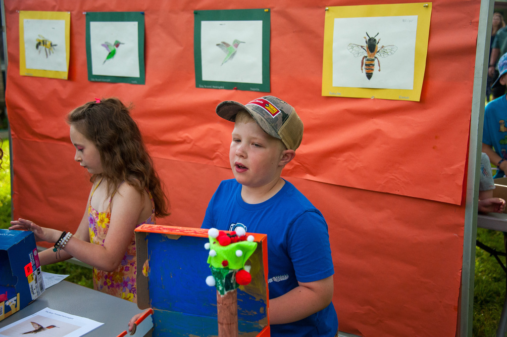 Two children look at artwork displayed on a table. One child holds a box with decorations. Behind them, orange fabric displays framed artwork.