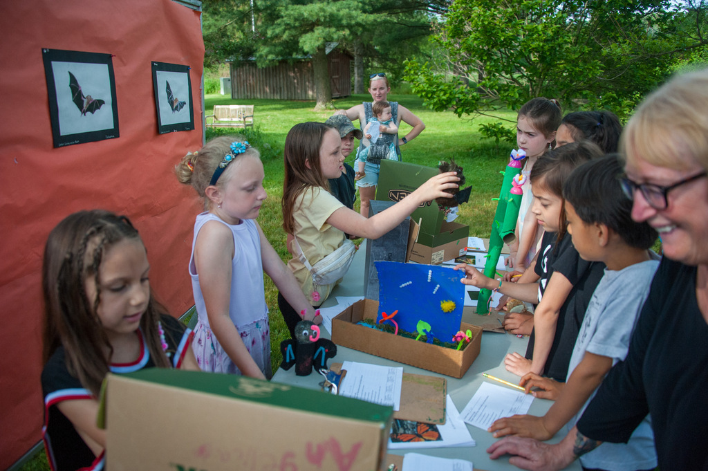 Kids and adults look at a table with crafts. A woman in glasses smiles at a child.