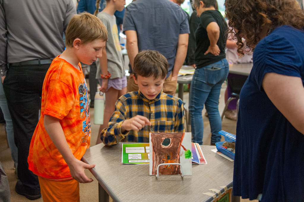 Three children gather around a table with an object. The boy in blue and yellow checks interacts with it.