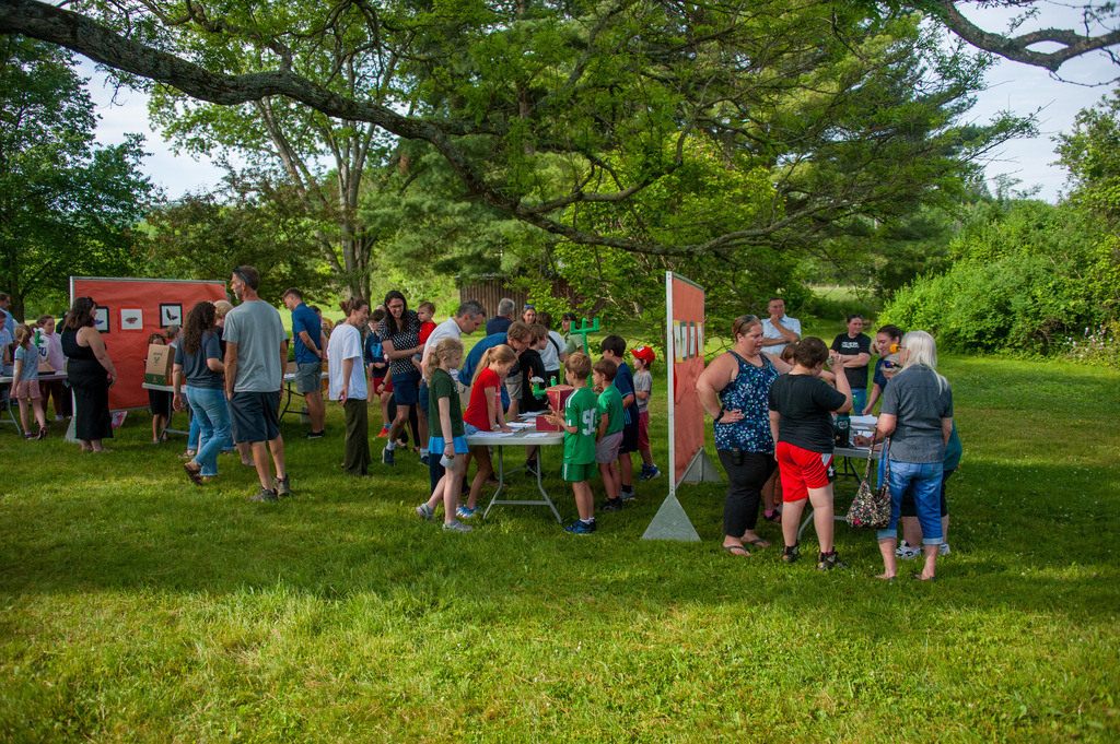 A group of people, some with hats, in a grassy field, some standing around tables with objects.