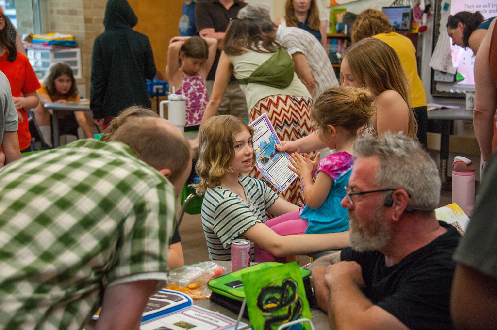 Adults and children gathered, some sitting, others standing, around a table; a girl holding a book.