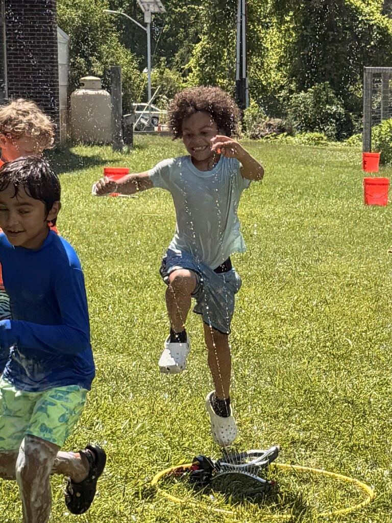 Children playing outside in grass. One child jumps over a hula hoop. Another child runs toward him.