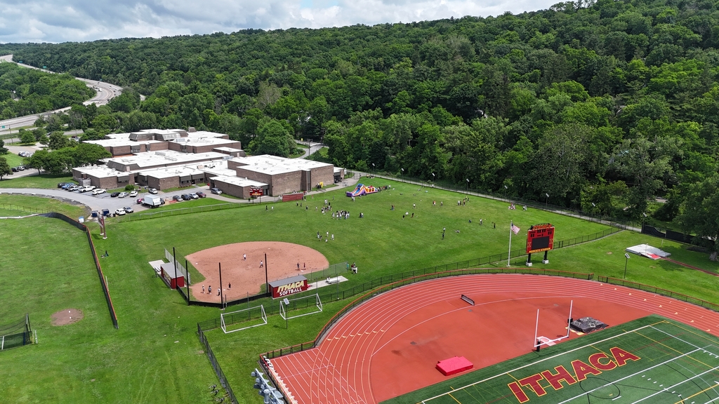 Aerial view of a sports facility with a red track, green field, and red bleachers. Buildings and trees surround the area.