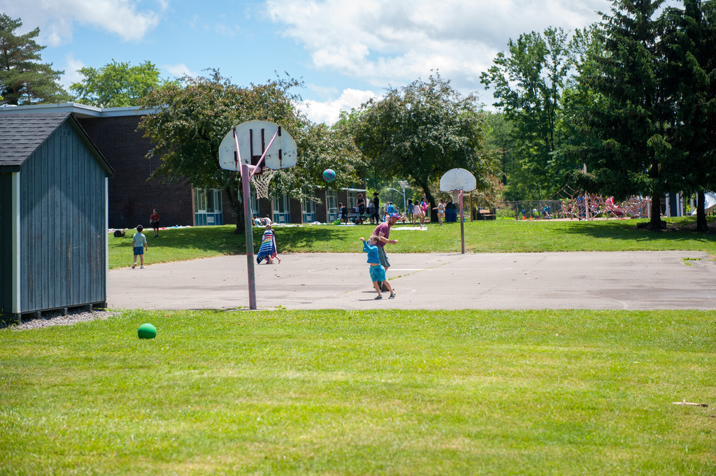 Outdoor playground with two basketball hoops and two children playing basketball in a grassy field.