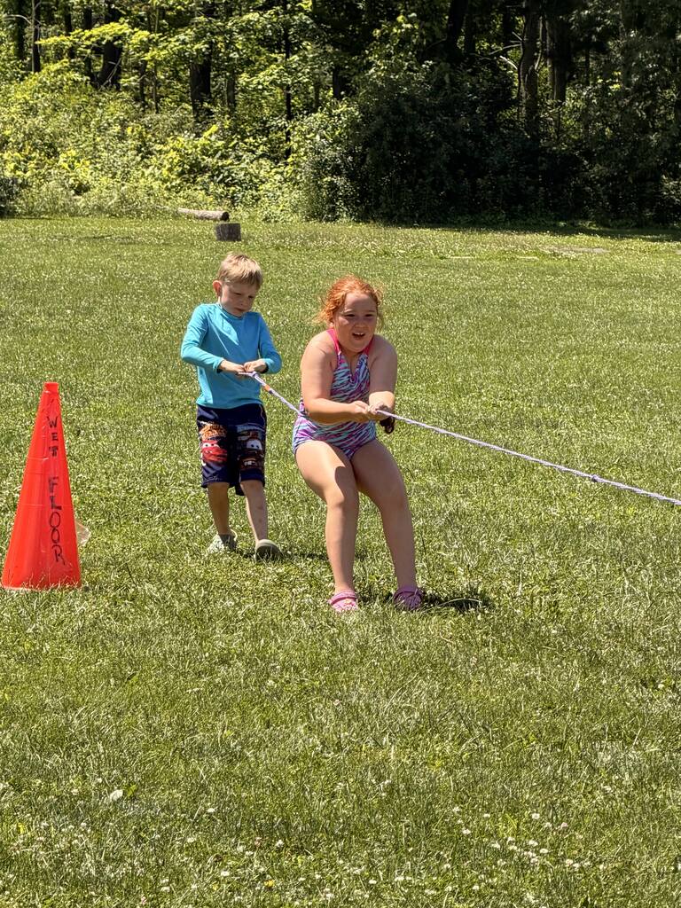 Two children pull on a rope with a traffic cone on the ground. Trees and a building are in the background.