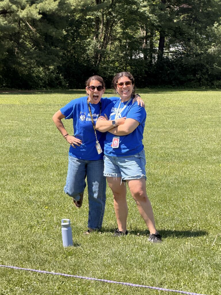 Two women in blue shirts stand in a grassy field with a small object on the ground. Trees are visible in the background.