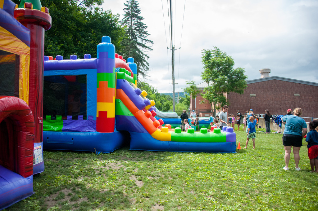 A grassy field with two colorful inflatables; one is a large slide. Children play near them, with an open building in the background.