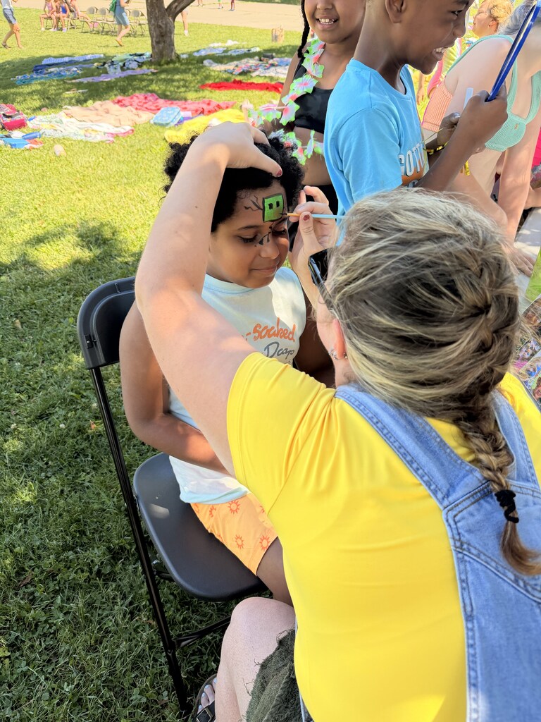 Woman in yellow shirt paints face art on a girl sitting on a lawn chair. Other people and items in background.