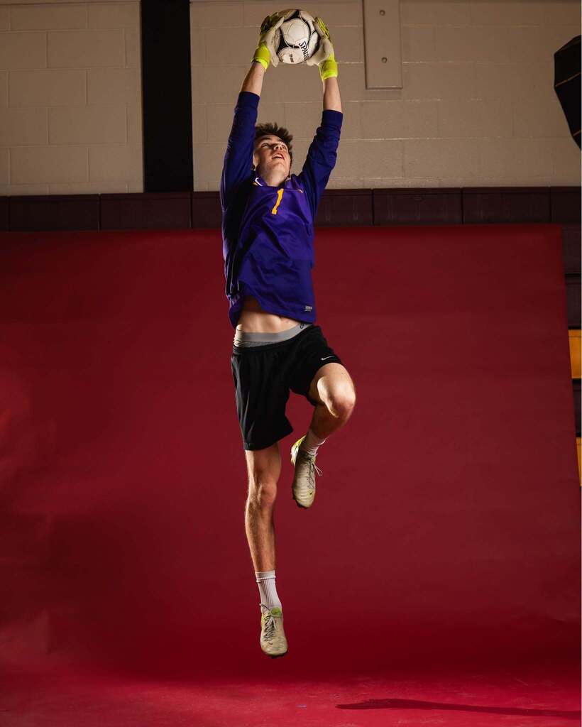 Person in blue long-sleeve shirt, black shorts, and white gloves jumps to catch a soccer ball.