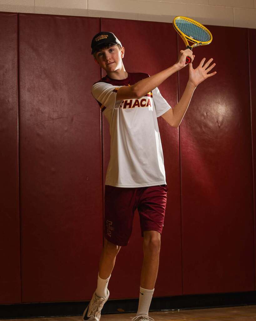 A young man in a white shirt with the letters HACA plays tennis, standing before a red wall.