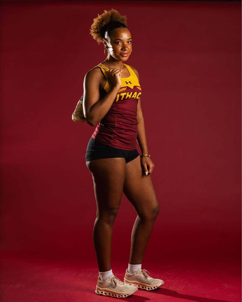 A woman in a maroon tank top and black shorts stands on a red background, holding a baseball bat.