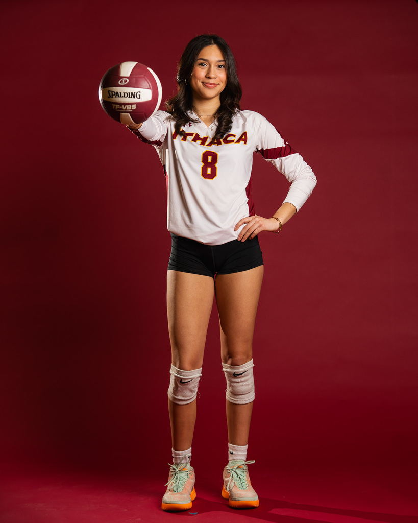 A volleyball player in a white uniform holds a ball. The player wears a shirt with "THACA" and the number 8.