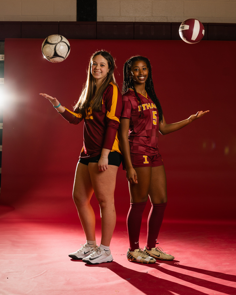 Two women in matching sports uniforms, one with a volleyball in the air. Red background.