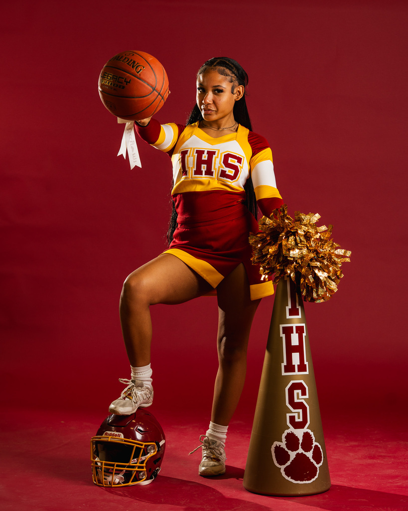 Person in red, yellow, and white cheerleading uniform holding a basketball and a pom-pom.