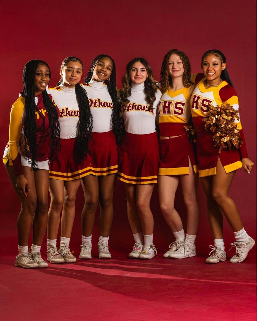 Five cheerleaders stand in a line. They wear red and white uniforms and hold pom-poms. They are on a red background.