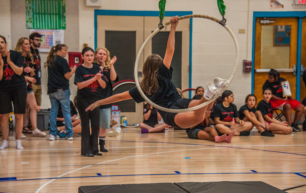 A woman performs a handstand on a hula hoop while others watch in a gymnasium.