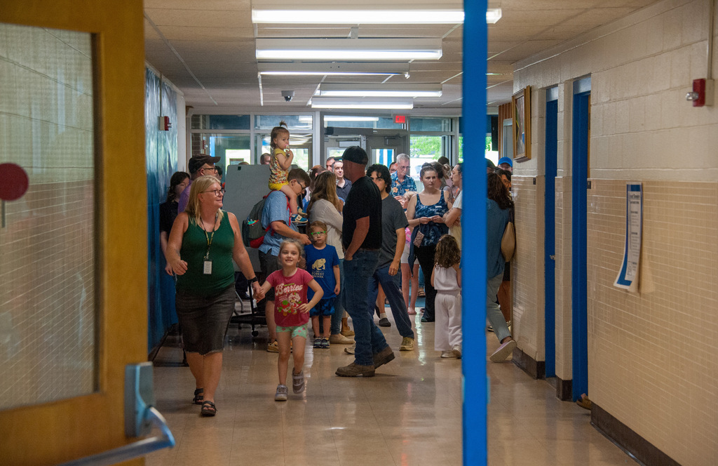 Many people, including a young child, walk in a school hallway with white walls and blue columns.