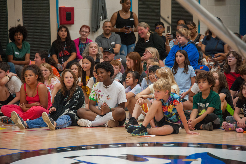 A large group of people sit on a basketball court in various positions, with a large logo on the floor.