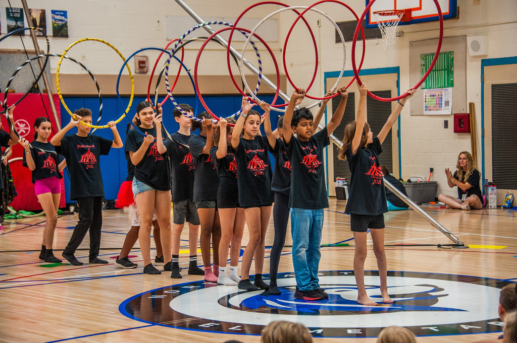 Kids hold up colorful hoops and stand in a circle, facing an audience in a gymnasium.