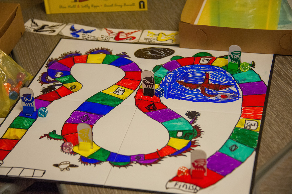 A colorful, hand-painted game board with a double-path layout, surrounded by game pieces and boxes on a wooden table.