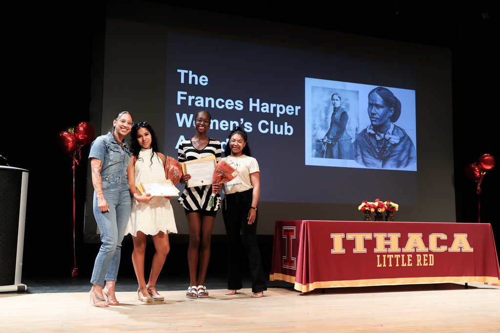Four people stand on stage in front of a screen displaying a woman's portrait. They hold certificates and stand beside a table with a red tablecloth.